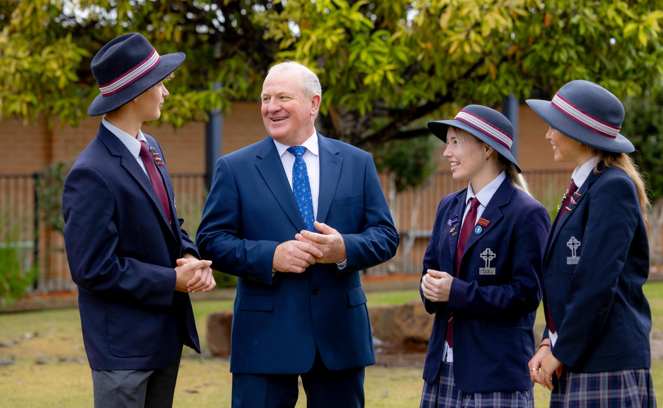 Students in school uniform blazers and hats smile and talk with a teacher outdoors